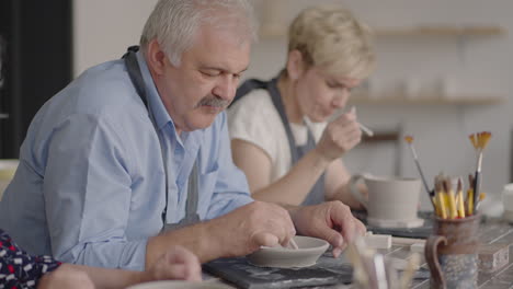 A-group-of-elderly-people-at-a-master-class-in-pottery-together-sculpt-and-cut-a-drawing-on-cups-of-clay-for-the-manufacture-of-ceramic-dishes